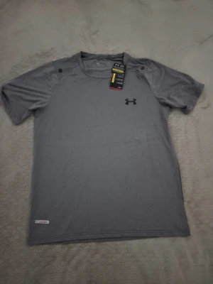 Grå tränings-tshirt från Under Armour - Snygg grå tränings-tshirt från Under Armour med korta ärmar och rund hals. Tillverkad i ett lätt och snabbtorkande material, perfekt för träning. Diskret logga på bröstet och meshstruktur för extra ventilation.