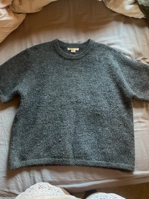 Mörkgrå stickad t-shirt - Mysig mörkgrå stickad t-shirt med korta ärmar och rund halsringning. Tröjan har en avslappnad passform och är tillverkad i ett mjukt, grovstickat material. Perfekt för lager-på-lager och enkel att styla till olika outfits.