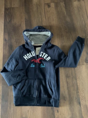 Mörkblå hoodie från Hollister - Mörkblå zip-hoodie från Hollister med broderad logga och texten 'So Cal' på bröstet. Hoodien har huva med grått foder, dragkedja framtill och två fickor. Perfekt för en avslappnad och cool stil.