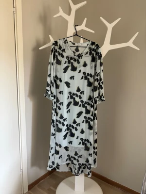 Långklänning med trumpetärm - Snygg långklänning i turkosa-mörkt blå toner med abstrakt mönster. Klänningen har en integrerad underklänning i bommullsjersey.  V-ringning, trumpetformade trekvartsärmar och är tillverkad i ett lätt, luftigt material som passar perfekt för varmare dagar. Den har en lös passform och faller fint längs kroppen.