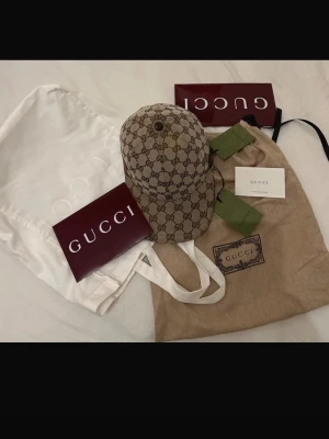 Gucci keps - Snygg keps från Gucci i beige canvas med klassiskt GG-monogram över hela kepsen. På sidan finns den ikoniska gröna och röda randen. Justerbar rem baktill. Perfekt accessoar för att lyfta din streetwear-look. Hör av er vid fler frågor priset kan såklart gå ner vid en affär 