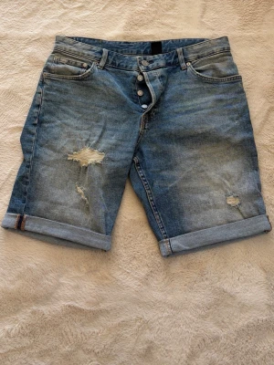 jeansshorts från &Denim - Snygga blå jeansshorts från &Denim i slim fit-modell. De har slitna detaljer, uppvikta benslut och klassisk femficksdesign. Perfekta för en avslappnad stil med lite edge.