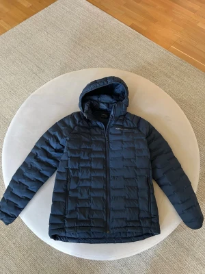 Mörkblå dunjacka från Peak Performance - Snygg mörkblå pufferjacka från Peak Performance med avtagbarhuva i junoirstorleken 170. Jackan har quiltad design, två sidofickor med dragkedja och diskret logga på bröstet. Perfekt för kyliga dagar och enkel att matcha med olika outfits.