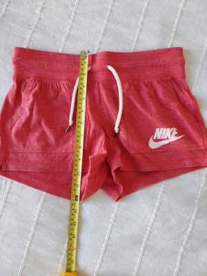 Rosa Nike shorts med snörning - Snygga shorts i korall från Nike med vit logga på ena benet och vit snörning i midjan. Modellen är kort och har två fickor framtill. Perfekta för sommarens alla aktiviteter.