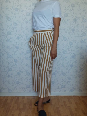 Randiga croppade byxor i beige och vitt - Säljer ett par croppade byxor med hög midja, faux ficka bak och raka ben. Byxorna har vertikala beige och vita ränder, bälteshällor och fickor fram. Perfekta för en trendig och avslappnad look.