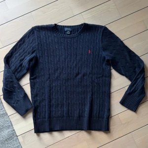 Mörkblå stickad tröja från Polo Ralph Lauren - Säljer en stilren mörkblå stickad tröja från Polo Ralph Lauren med kabelstickat mönster. Tröjan har en rund halsringning och ett diskret rött logotypbroderi på bröstet. Perfekt för kyligare dagar!