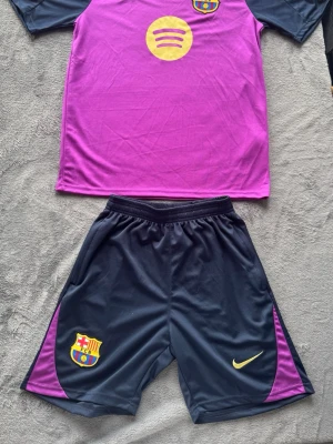 Barcelona kit - Säljer ett FC Barcelona träningsset från Nike med lila tröja och marin blåa shorts. Tröjan har klubbmärke, Nike-logga och Spotify-tryck på bröstet. Shortsen är svarta med lila detaljer och klubbmärke samt Nike-logga. Perfekt för fotbollsträning eller match.