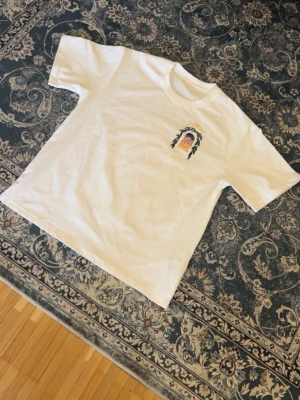 Vit t-shirt Avenida Casablanca - Vit t-shirt med tryck på bröstet som föreställer en portal med texten 'Avenida Casablanca' och ett färgglatt motiv. T-shirten har klassisk passform och rund hals. Perfekt för dig som gillar stilrena plagg med unik detalj.