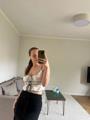 Vit topp med snörning - Trendig vit croppad topp med smala axelband och snygg snörning framtill. Toppen har volangdetaljer och en feminin v-ringning. Perfekt att styla med höga byxor eller kjol för en cool look.