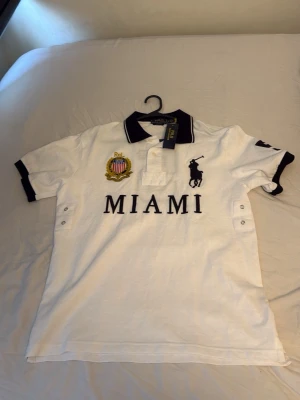 Vit pikéskjorta från Ralph Lauren - Vit pikéskjorta från Ralph Lauren med svart krage och detaljer. Stor broderad logga på bröstet, texten MIAMI och emblem. Kortärmad modell i bomull med knappar vid sidorna och klassisk polokrage. Helt ny