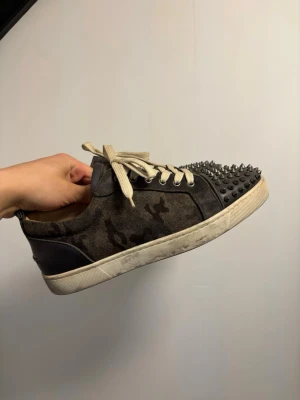 Christian Louboutin jr spike camo - Unika sneakers i camomönstrad mocka och svart läder från Christian Louboutin. Skorna har vita snören och coola svarta nitar på tån för en edgy look. Perfekt för dig som vill sticka ut med din stil.