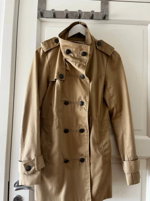 Beige trenchcoat från ONLY - Stilren beige trenchcoat från ONLY med dubbelknäppning, axelklaffar och bred krage. Jackan har svarta knappar, fickor i sidorna och justerbara remmar vid ärmsluten. Perfekt för dig som gillar klassisk och tidlös stil.