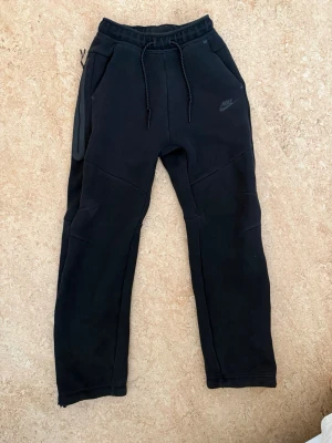 Svarta Nike tech mjukisbyxor med dragsko - Svarta mjukisbyxor från Nike med dragsko i midjan och två sidofickor. Byxorna har en sportig look med diskret Nike-logga på låret och paneldetaljer vid knäna. Tillverkade i mjukt och bekvämt material, perfekta för chill eller träning.