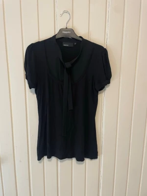 Svart blus med knyt från KappAhl - Stilren svart blus från KappAhl med korta puffärmar och dekorativ knytning i halsen. Blusen har en mjuk och följsam passform, perfekt för en trendig och enkel look. Tillverkad i ett lätt material som känns skönt mot huden.
