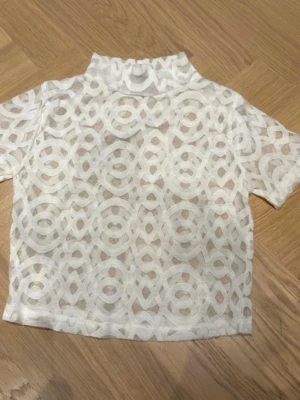 Vit mesh polotopp från ASOS - Snygg vit mesh polotopp från ASOS med korta ärmar och ett unikt geometriskt mönster. Toppen är genomskinlig och har en hög krage som ger en trendig look. Perfekt att styla lager på lager eller med ett linne under.