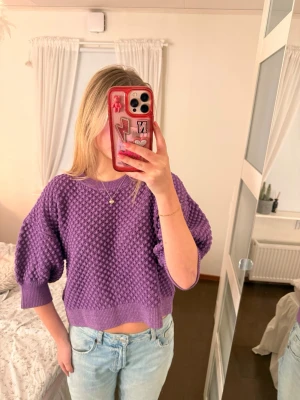 Lila stickad tröja med puffärm - Trendig lila stickad tröja med kortare passform och puffiga trekvartsärmar. Tröjan har ett strukturerat mönster och breda ribbade muddar vid ärmslut och nederkant. Perfekt att styla med jeans för en cool vardagslook.