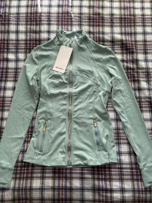 Lululemon luon define jacket i fog green gold zip - Supersnygg ljusgrön träningsjacka från Lululemon med figurnära passform, hel dragkedja framtill och två fickor med dragkedja. Tillverkad i ett stretchigt funktionsmaterial som andas. Perfekt för träning eller som ett extra lager på väg till gymmet. Storlek 4 eller XS (passar S också)