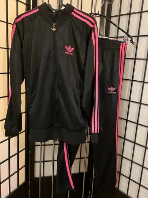adidas Originals Y2K tracksuit set 🖤💖 -  Svart set med hot pink stripes + trefoil logos. Ger exakt mcbling / Bratz / 2000s it-girl vibe ✨ Jackan sitter relaxed medan byxorna är mer slim/cropped vilket ger sjukt snygg silhouette on body.  XS-ish fit 🌸  Mått ca: Jacka p2p 49–50 cm / längd 60–61 cm   Byxor waist 27–28 cm (stretch 38–40) / längd 86 cm / inseam 61–62 cm  Väldigt fint vintage-skick.