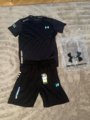Svart Under Armour - Snyggt svart träningsset från Under Armour med t-shirt och shorts. T-shirten har tryck i neonfärger och logga på bröstet, medan shortsen har elastisk midja och färgglada detaljer längs sidan. Perfekt för gymmet eller löpning. Materialet är lätt och andas.