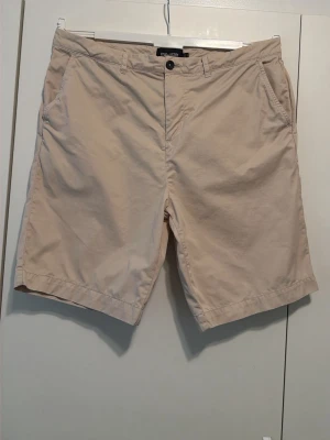 Beige shorts från Lyle & Scott - Snygga beige shorts från Lyle & Scott med klassisk passform och diskret logga bak. Tillverkade i mjuk bomull, har midwaist, sidofickor och bakfickor med knappar. Perfekta för sommaren och enkel att matcha med olika stilar.