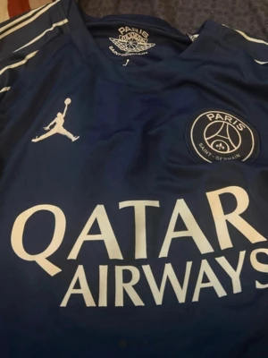 PSG matchtröja blå Jordan - Snygg blå Paris Saint-Germain matchtröja med vita detaljer och Qatar Airways-tryck på bröstet. PSG-logga och Jordan-logga på framsidan, samt namn och nummer 14 (D. Doué) på ryggen. Tillverkad i lätt och ventilerande material, perfekt för fotboll eller träning.