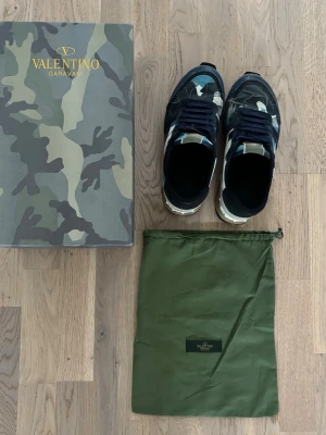 Valentino Garavani camo sneakers - Snygga sneakers från Valentino Garavani med unik camo-design i blått, svart, vitt och grått. Skorna är smutsiga och mycket synligt använt. Storlek 41. Kommer med Dustbag och box! Priset är ej hugget i sten den har hål men går att fixa!