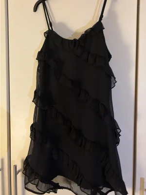 Svart volangklänning från Gina Tricot - Svart kort klänning med tunna axelband och flera volangdetaljer i diagonala linjer. Klänningen har en luftig och lätt känsla, perfekt för dig som gillar en feminin och trendig stil. Materialet är troligtvis polyester och modellen är ärmlös.