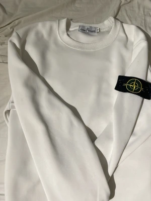 Vit sweatshirt från Stone Island - Säljer en clean vit sweatshirt från Stone Island med klassisk logga på ärmen. Tröjan har rund halsringning och är gjord i mjukt bomullsmaterial. Perfekt för dig som gillar streetwear och vill ha en stilren look.