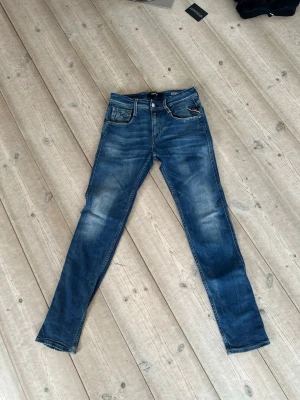 Blå smala jeans från Replay - Snygga blå jeans från Replay med smal passform och klassisk femficksdesign. Jeansen har slitningar och ljusare partier framtill för en cool, använd look. Tillverkade i denim med detaljerade sömmar och läderpatch bak i midjan. Storlek 28x32 replay Anbass slim fit 