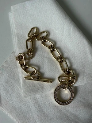 Chunky guldarmband från SNÖ - Stilrent chunky armband i guldfärgad metall med stora länkar och ett cirkelformat hänge dekorerat med små glittrande stenar. Armbandet har en snygg T-lås detalj som gör det enkelt att ta på och av. Från SNÖ