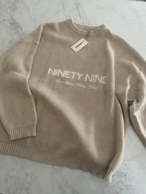 Beige stickad tröja NINETY NINE - Beige stickad tröja med texten 'NINETY NINE' i vitt framtill. Tröjan har rund halsringning, ribbade muddar och avslut. Perfekt för en avslappnad och trendig look.
