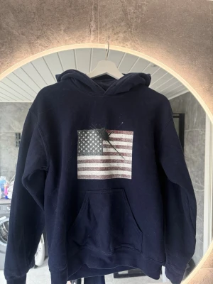 Mörkblå hoodie med USA-flagga - Mörkblå hoodie med stor ficka framtill och tryck av amerikanska flaggan på bröstet. Flaggan har en cool färgfläck över sig för en unik look. Hoodien har huva och ribbade muddar. Perfekt för dig som gillar streetwear och vill sticka ut. Ej tvättad på bild han aldrig tvätta den. Skriv vid de minsta fundering// William