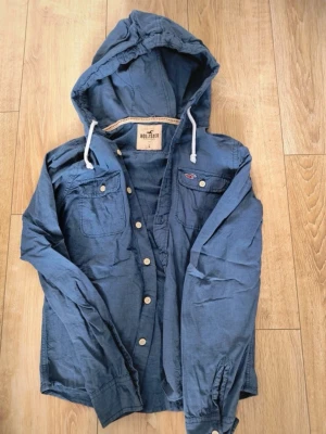 Blå overshirt jacka från Hollister - Snygg blå overshirt jacka från Hollister med huva och dragsko. Jackan har knappar framtill, två bröstfickor med lock och långa ärmar. Perfekt för lager-på-lager och en avslappnad stil. Står som L men skulle snarare beskriva som S/M
