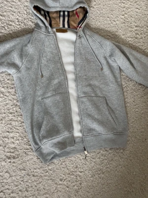 Burberry zip hoodie grå  - Säljer en snygg och stilren Burberry zip hoodie i ljusgrå färg med klassiskt rutigt foder i huvan. Mjuk och bekväm passform med dragkedja framtill och två fickor. Diskret broderad Burberry-logga på bröstet.