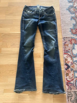 Bootcut jeans från True Religion - Mörkblå bootcut jeans från True Religion med vita kontrastsömmar och klassiska broderade bakfickor. Jeansen har låg midja och slitna detaljer vid benslut och lår. Tillverkade i denim med stretch för extra komfort.