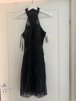 Svart glittrig halterneckklänning - Svart kortklänning med halterneck och glittriga paljettdetaljer och pärlor i ett elegant mönster. Klänningen har en figurnära passform upptill och lätt utställd kjol. Perfekt för dig som vill sticka ut med en snygg och trendig look. Klänningen är även öppen i ryggen. Klänningen är i nyskick med prislapp på med ett ursprungspris på 4500.