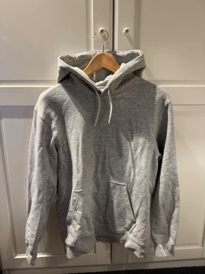 Grå basic hoodie med ficka - En stilren grå hoodie med huva och dragsnören. Hoodien har en klassisk känguruficka framtill och ribbade muddar vid ärmslut och nederkant. Perfekt för en avslappnad och trendig look. Materialet känns mjukt och bekvämt mot huden.