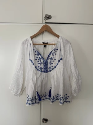 Vit boho-blus med blå broderier - Vit bohemisk blus med blå broderade detaljer och tofsar. Blusen har v-ringning med knytband och trekvartsärm. Tillverkad i ett lätt, krinklat material som ger en avslappnad look. Perfekt för en chill och trendig stil.