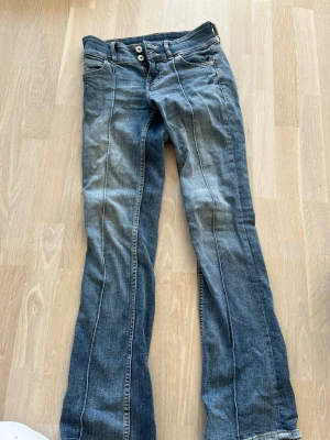 Low waist bootcut jeans - Snygga blå bootcut jeans med dubbla knappar i midjan och klassiska fem fickor. Jeansen har ljusa slitningar och markerade sömmar längs benen för en cool look. Perfekta för dig som gillar en avslappnad men trendig stil.