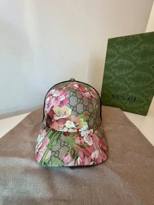 Gucci keps med blommigt mönster - Snygg keps från Gucci med klassiskt GG-mönster i beige och brunt, täckt av färgglada blommor i rosa, vitt och grönt. En mycket fin keps perfekt till det varma sommarvädret och till din outfit.
