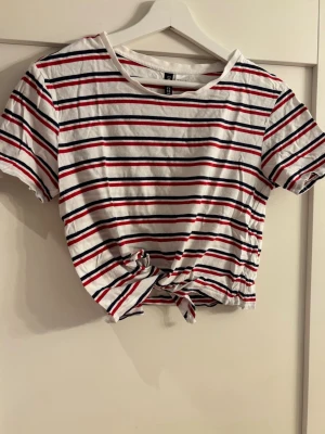 Randig croppad topp från H&M - Säljer en croppad topp från H&M med vita, röda och marinblå ränder. Toppen har korta ärmar, rund halsringning och knyt framtill för en avslappnad vibe. Perfekt till jeans eller kjol för en fräsch look.