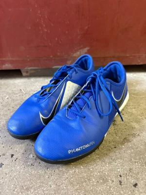 Nike Phantom Vision blå fotbollsskor - Säljer ett par blå Nike Phantom Vision fotbollsskor med vita och svarta detaljer. Skorna har snörning, låg profil och ovandel i syntetmaterial för bra bollkontroll. Snygg design med Nike-logga på sidan och texten PHANTOM VSN på tåpartiet.
