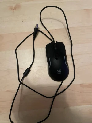 Svart SG gamingmus med USB-kabel - En svart gamingmus från SG med USB-anslutning och lång kabel. Musen har ergonomisk form, två sidoknappar och scrollhjul. Passar bra till gaming och datoranvändning. Tillverkad i plast med detaljer i svart och grått.