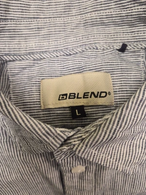 Randig skjorta från Blend, storlek L - Snygg vit och blå randig skjorta från Blend i storlek L. Skjortan har klassisk krage, lång ärm och bröstficka. Perfekt för en stilren look och enkel att matcha med jeans eller chinos.