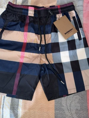 Rutig Burberry shorts med snörning - Snygga shorts från Burberry med klassiskt rutigt mönster i marinblå, beige, svart, vit och röda toner. Shortsen har elastisk midja med snörning och sidofickor. Perfekta för sommaren och ger en trendig look.