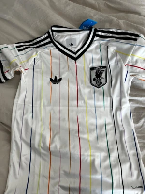 Japan VM Tröja 2026 Borta - Snygg vit Japan fotbollströja från Adidas med svart-vita detaljer och JFA-logga på bröstet. Tröjan har V-ringning, korta ärmar och är tillverkad i lätt, ventilerande Climalite-material. Perfekt för fotboll eller träning.