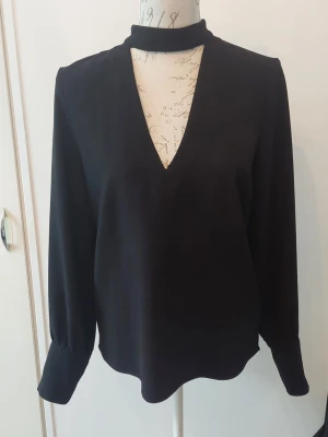 Svart blus med djup V-ringning - Stilren svart blus med lång ärm och hög krage. Blusen har en djup V-ringning framtill och vida ärmslut som ger en trendig look. 