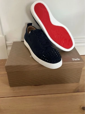 Blå Christian Louboutin sneakers - Snygga blå sneakers från Christian Louboutin med klassiska röda sulor och svarta nitar på tån. Skorna har en låg profil, snörning och är tillverkade i mocka. Perfekt för dig som vill sticka ut med en edgy och exklusiv look.