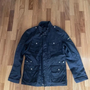 Woolrich jacka  - Wool rich jacka  med flera fickor och knappar framtill. Jackan har en hög krage och långa ärmar, perfekt för kyligare dagar. Praktisk och stilren design. Storlek S