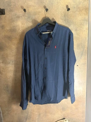 Marinblå Lin skjorta från Polo Ralph Lauren - Stilren marinblå skjorta från Polo Ralph Lauren med klassisk krage och knappar framtill. Skjortan har lång ärm och ikonisk röd logga broderad på bröstet. Perfekt för en snygg och avslappnad look.